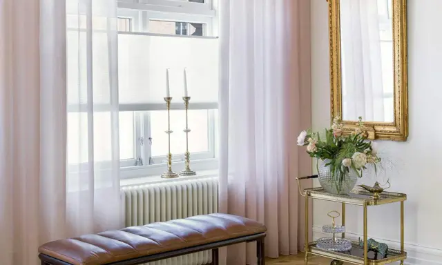 Ljusrosa gardiner längd från golvet ramar in ett fönster med ljusinsläpp. En brun läderbänk står framför ett element.