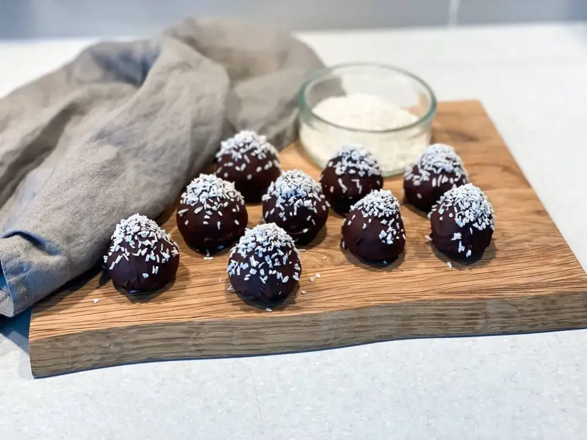 Mörka, chokladöverdragna protein chokladbollar toppade med kokosflingor ligger på en träskärbräda.
