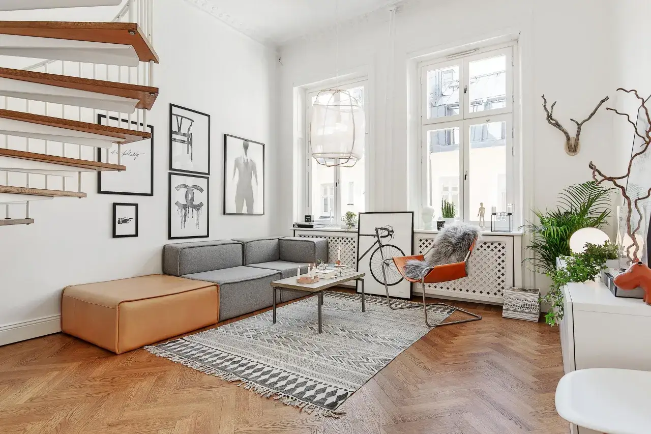 Vardagsrum med grå soffa, orange fåtölj, trägolv och en trappa. En stor matta med mönster och konst på väggarna bidrar till den klassiska designen.