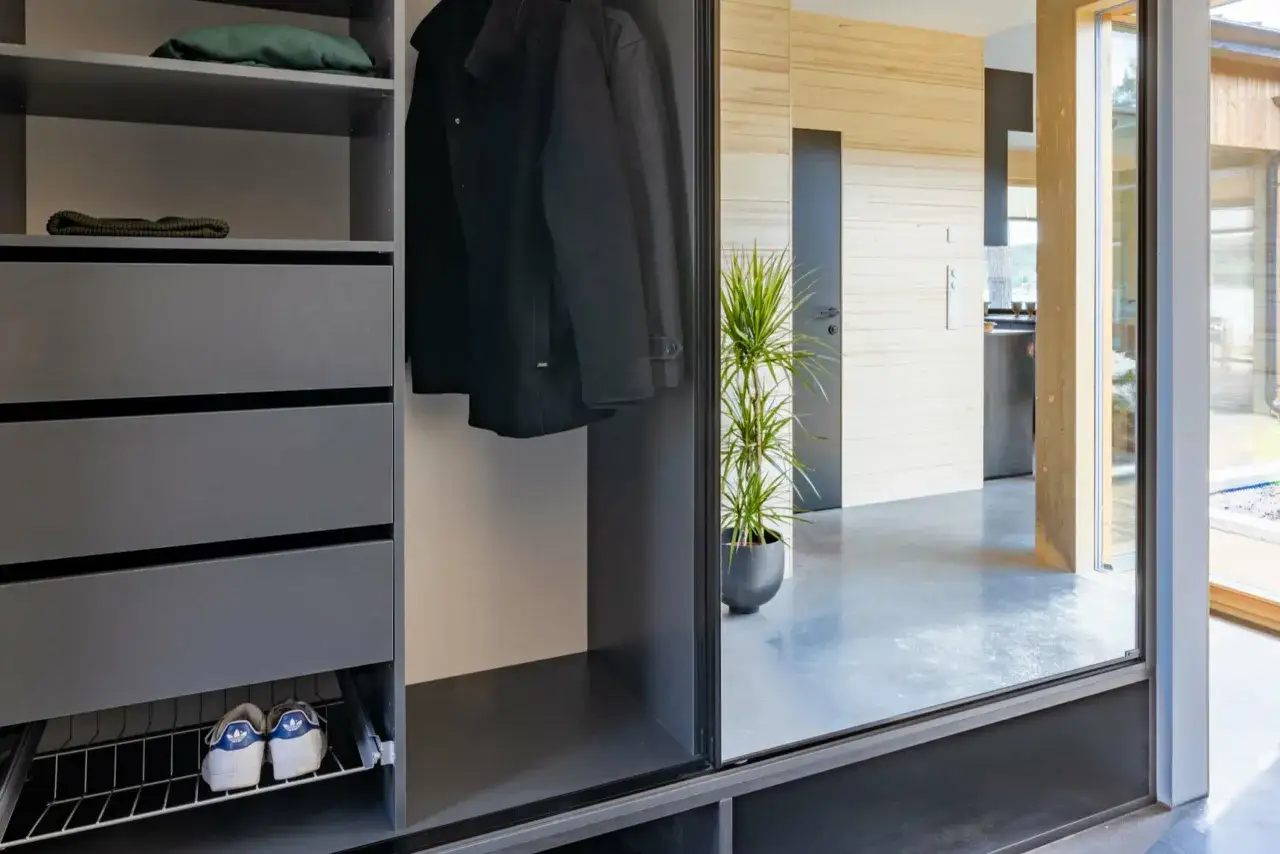 En modern garderobslösning med mörka lådor, hängande rockar och ett par sneakers. Spegeldörren reflekterar en hall med träpaneler och en grön växt.