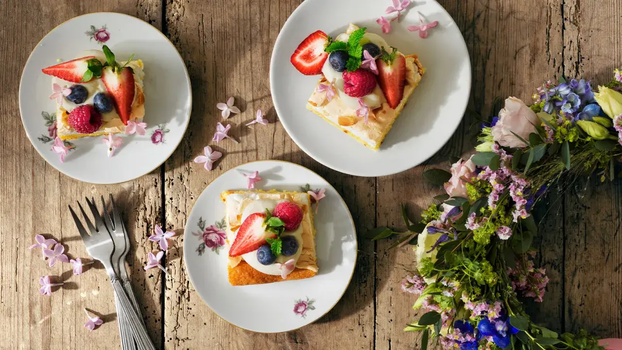 Tre tårtbitar med bär och grädde, perfekt vegetarisk midsommarmat. Blommor och gafflar på träbord.