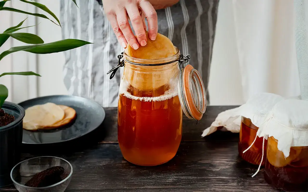 En hand placerar en SCOBY i en glasburk med fermenterad dryck. Ingredienser för kombucha recept syns på bordet.
