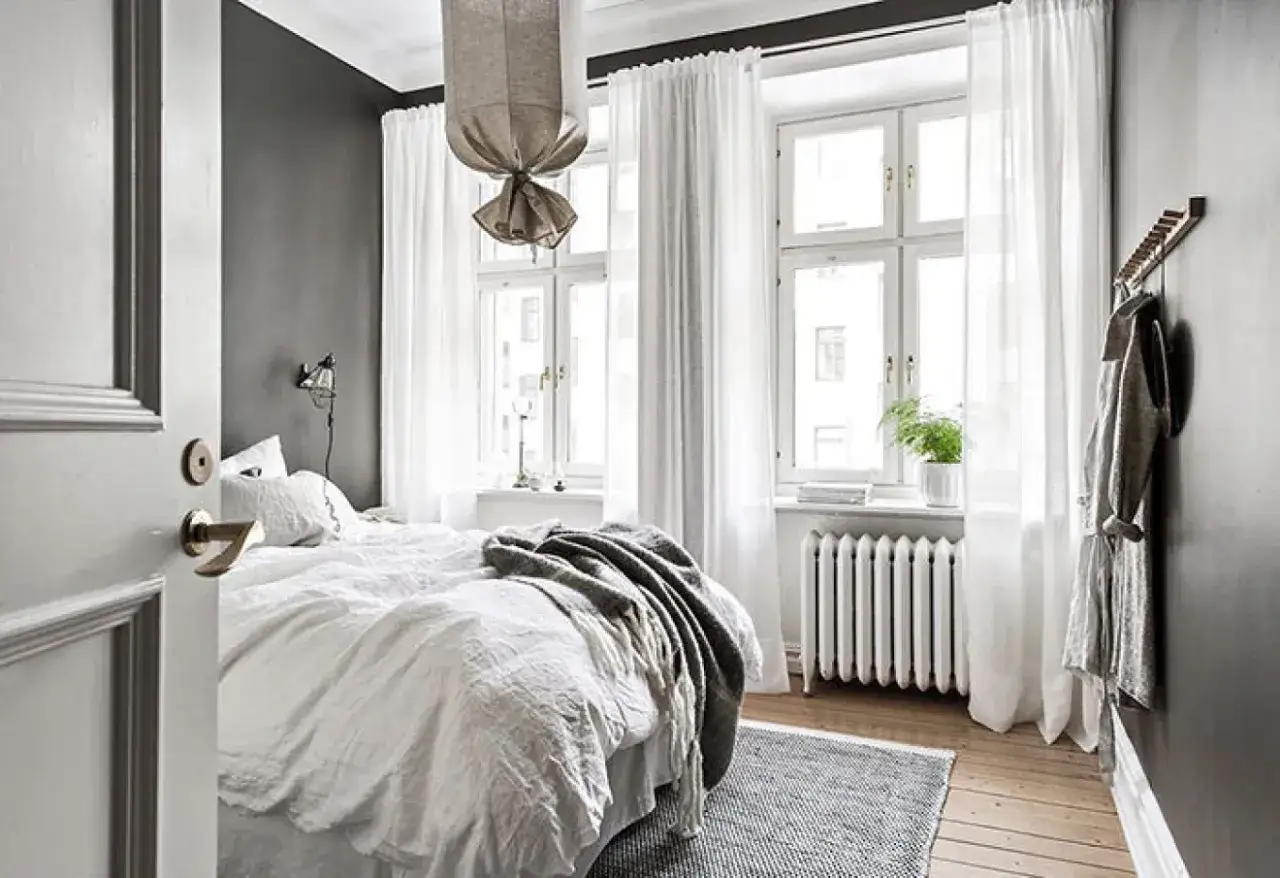 Ett sovrum med en stor, vit säng, en lång gardinstång med vita gardiner och en grå vägg.