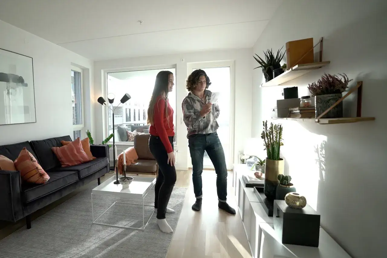 Två personer diskuterar något i ett modernt vardagsrum med trendig inredning, mörk soffa och hyllor med växter.