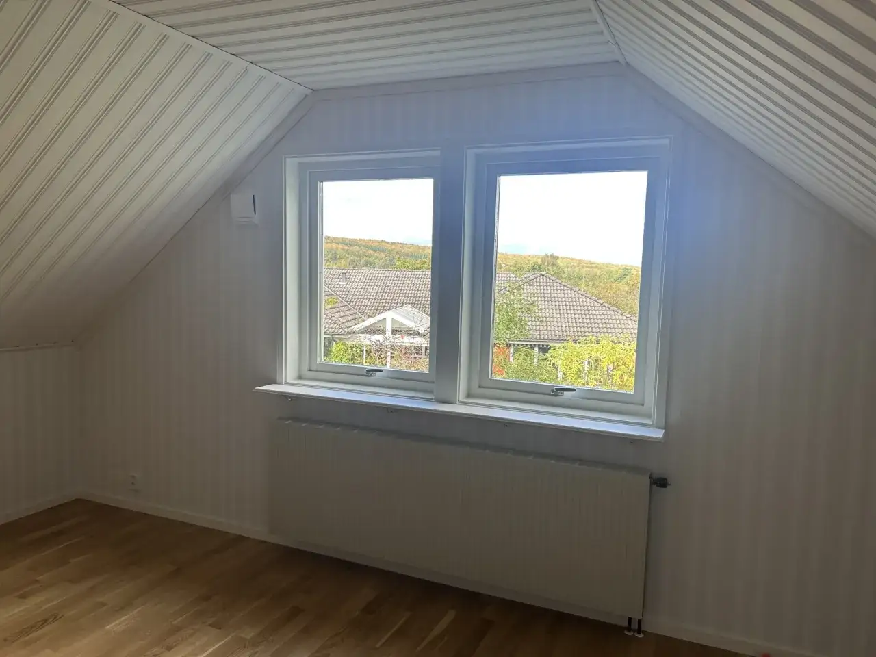 Vitt snedtak med randiga tapeter och ett fönster med utsikt över träd. Gardiner saknas, men ett element finns under fönstret.