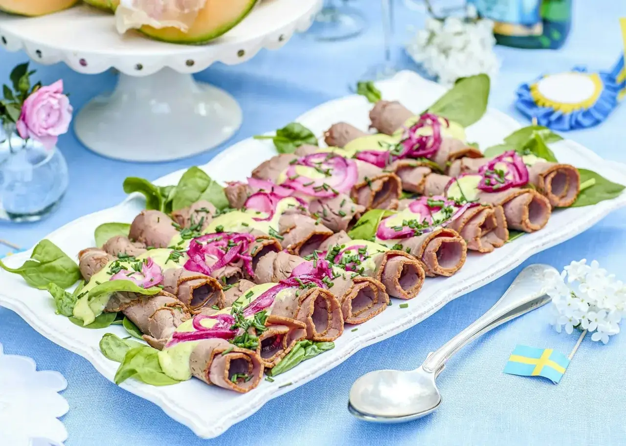 Rullad rostbiff med grön sås och rödlök, perfekt för ett somrigt buffé recept. Serveras på en vit bricka med gröna blad.