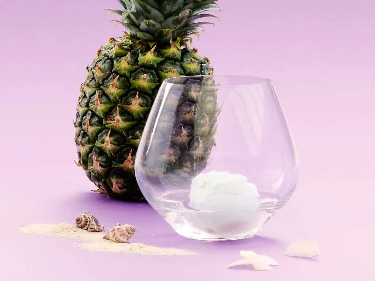En ananas står bredvid ett glas med en skopa glass, som förberedelse för en pina colada. Små snäckor och sand antyder en strand.