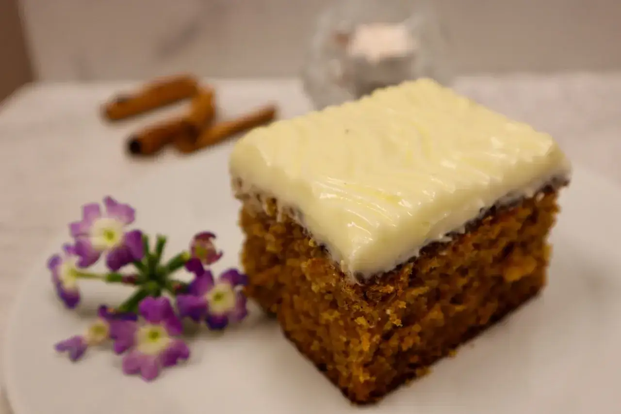 En bit glutenfri morotskaka med krämig frosting, dekorerad med lila blommor och kanelstänger i bakgrunden.