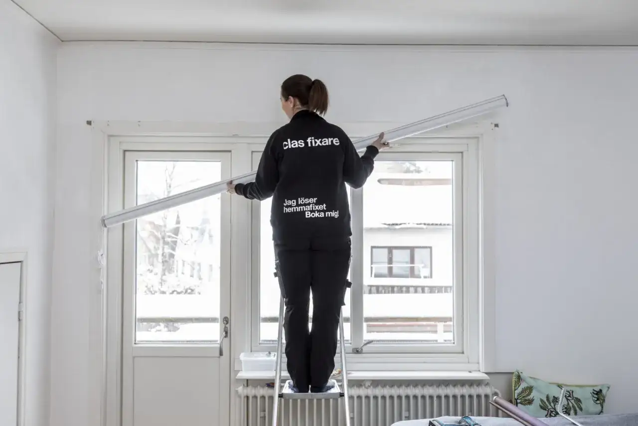 En person p&aring; en stege monterar en l&aring;ng gardinst&aring;ng v&auml;gg till v&auml;gg &ouml;ver ett f&ouml;nster.