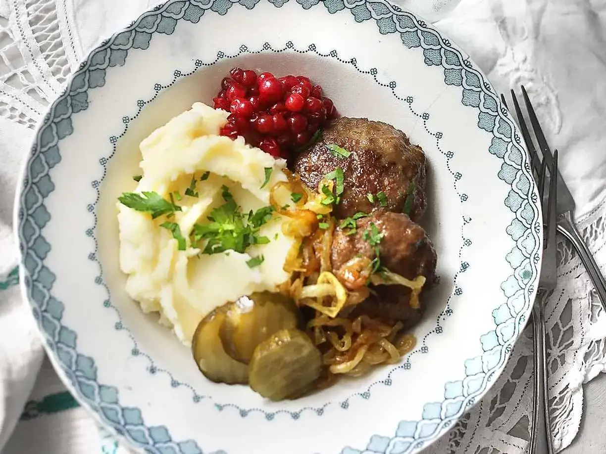 En tallrik med pannbiff i ugn, potatismos, lingon, stekt lök och saltgurka.