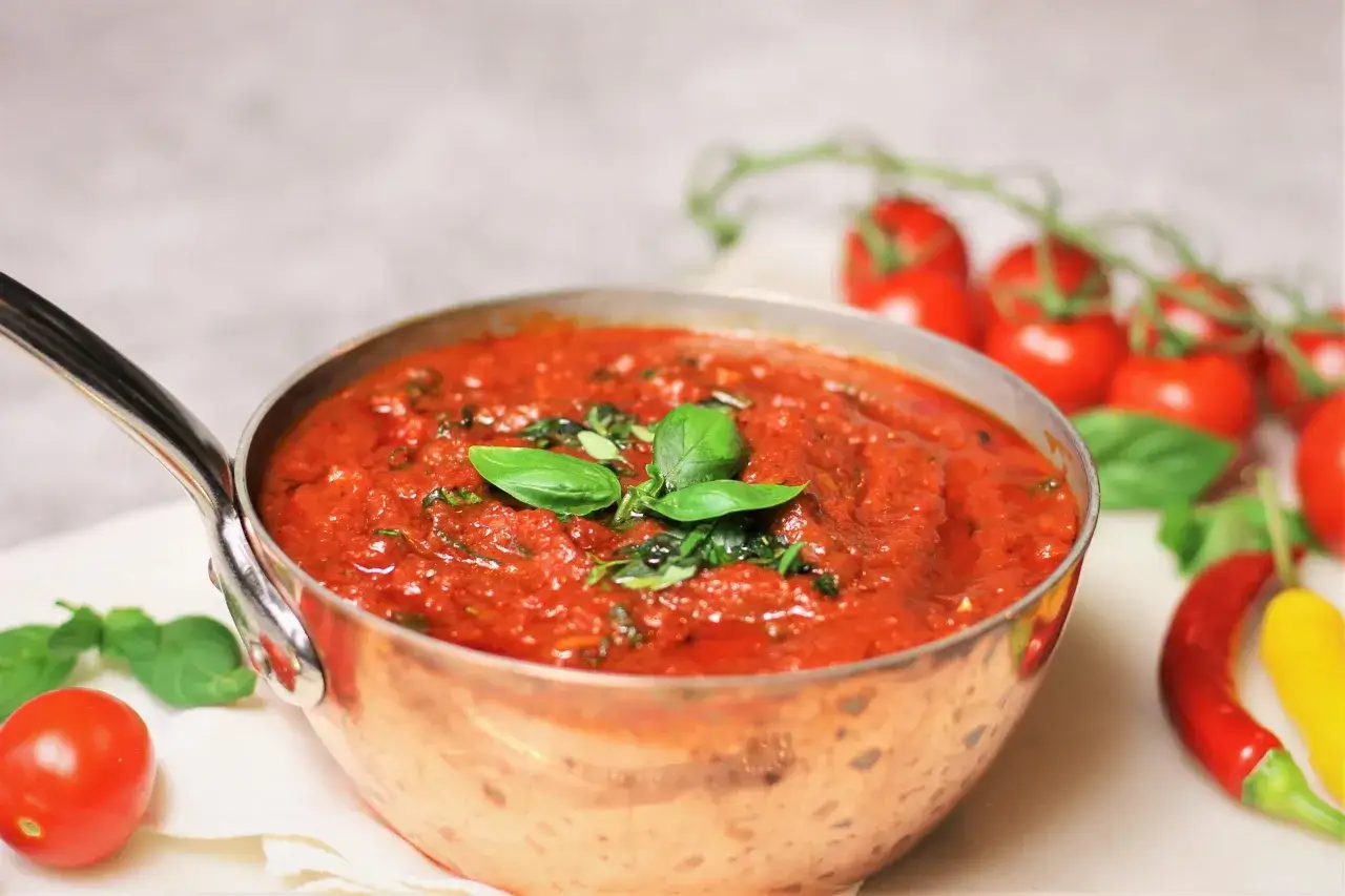 En kopp med hemlagad tomatsås, garnerad med färsk basilika. Bredvid syns tomater och chili, som ingredienser till ett gott tomatsås recept.