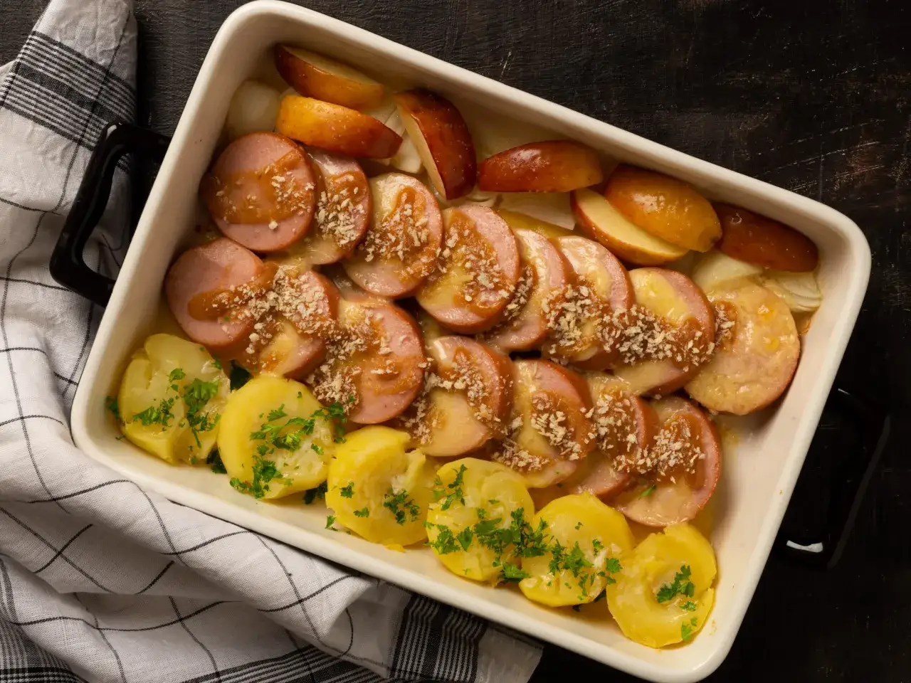 En ugnsform med korv i ugn, potatis, äpple och lök. Ströbröd och sås toppar korven.