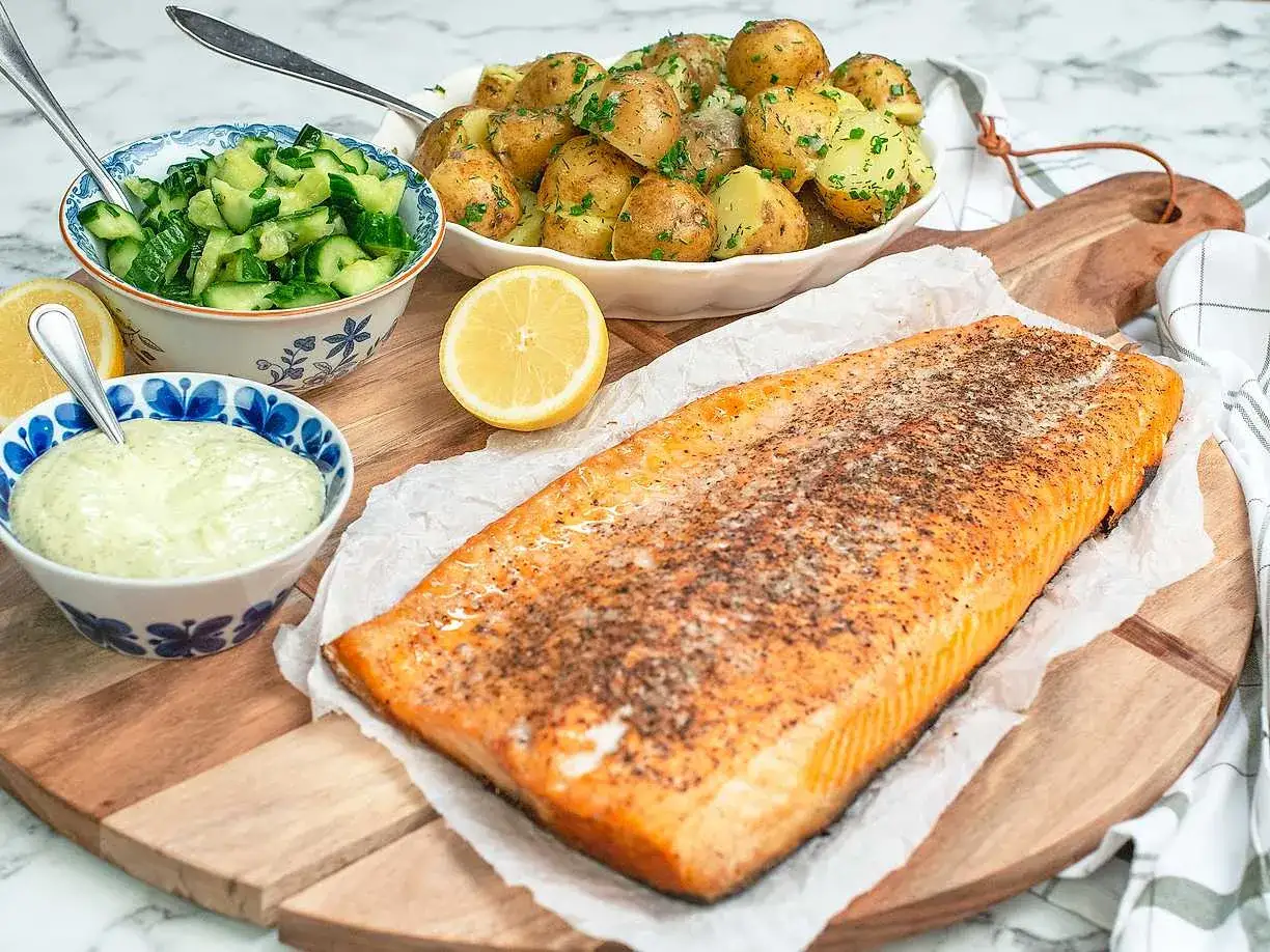 En saftig laxfilé, perfekt för att grilla lax, serveras med färskpotatis, gurksallad och en krämig sås.