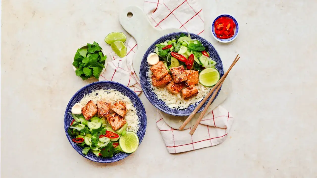 Två skålar med ris, teriyaki lax, gurka, chili och koriander. En liten skål med chiliskivor och limeklyftor.