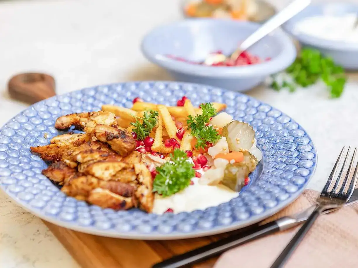 En tallrik med saftig kyckling kebab, pommes frites, picklad grönsaksmix, granatäppelkärnor och en krämig sås.