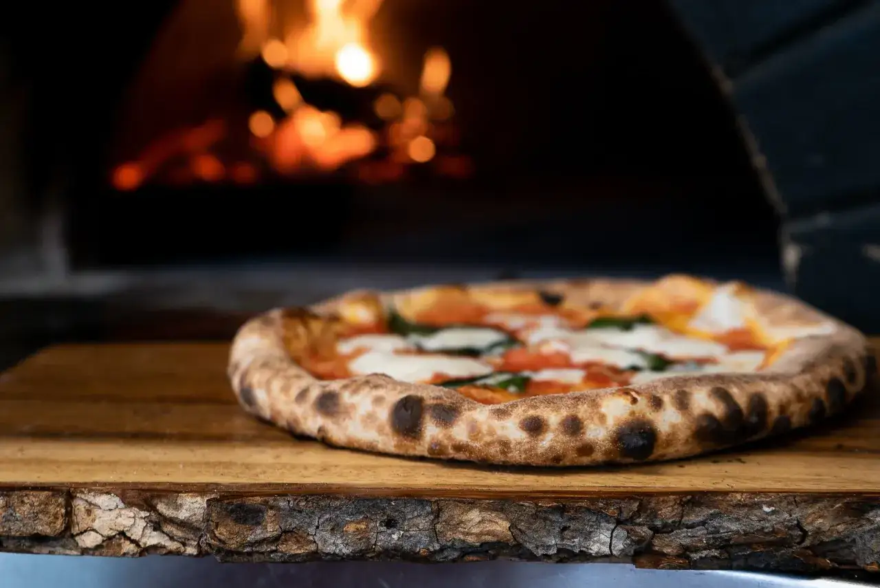 En nybakad, hemmagjord pizza med bubblande ost och tomater vilar på ett träfat framför en glödande pizzaugn.