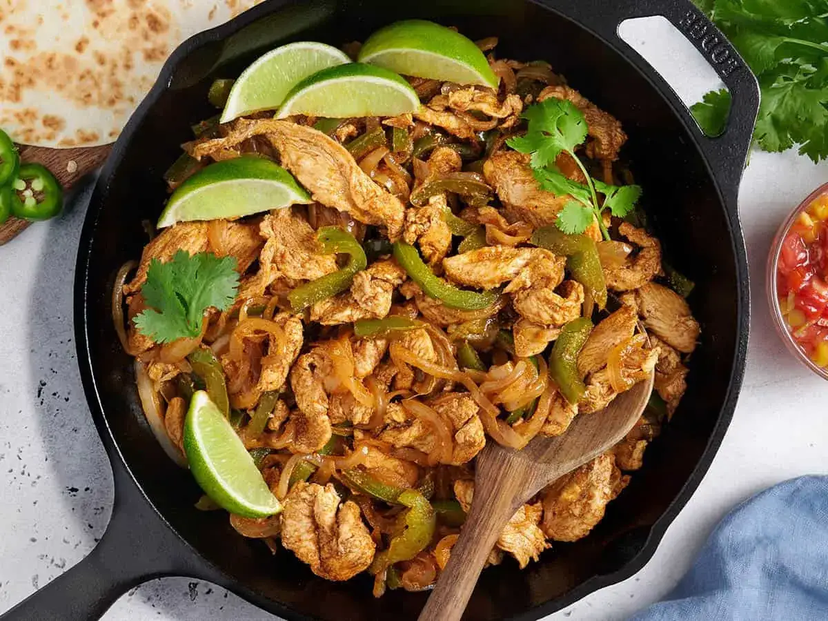 En gjutjärnspanna fylld med kyckling, paprika och lök, perfekt för ett gott fajitas recept. Garnerad med limeklyftor och koriander.