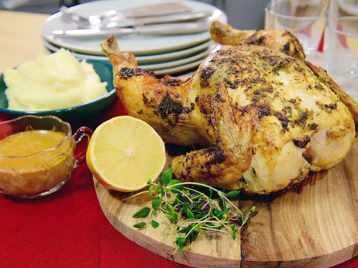 En saftig hel kyckling i ugn, serverad med krämigt potatismos, en skål med gyllene sås, en citronklyfta och färska örter.