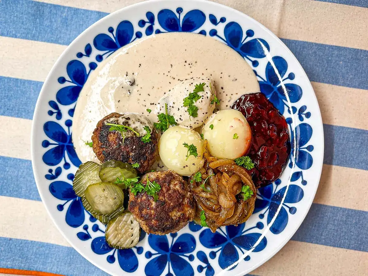 En tallrik med pannbiff, gräddsås, lingonsylt, pressgurka och kokt potatis. Ett klassiskt svenskt pannbiff recept.