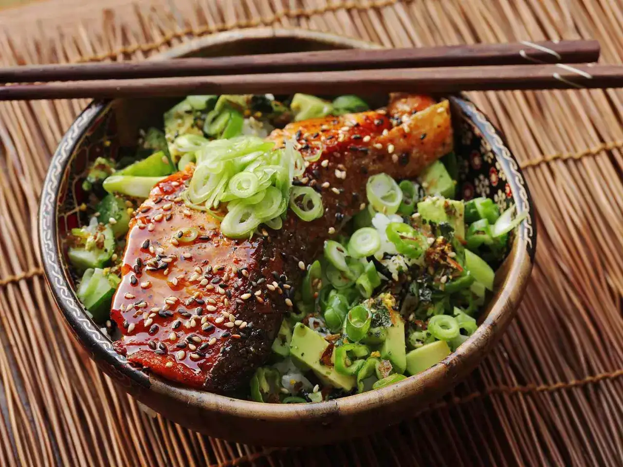 En skål med teriyaki lax, ris, avokado och salladslök, toppad med sesamfrön.