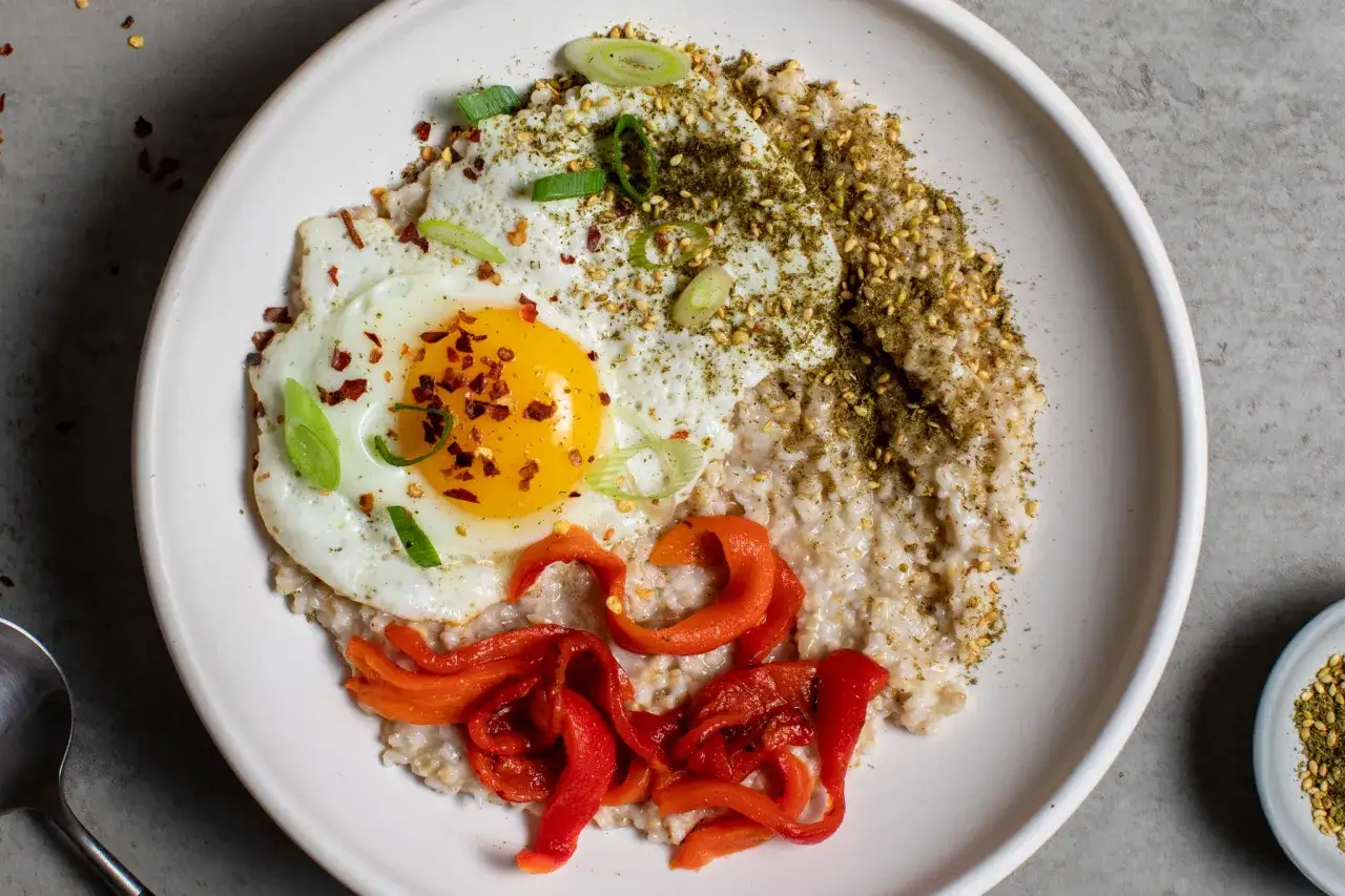 En tallrik med krämiga scrambled oats, toppad med ett stekt ägg, strimlad röd paprika, salladslök och kryddor.