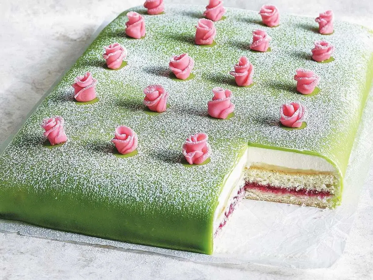 Grön tårtbotten i långpanna med rosa marsipanrosor och pudersocker. En bit är bortskuren och visar lager av grädde och sylt.