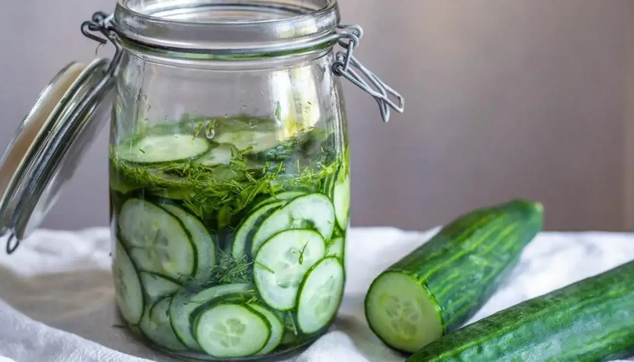 Burk med skivade gurkor och dill, redo för ett enkelt saltgurka recept. Två färska gurkor ligger bredvid.