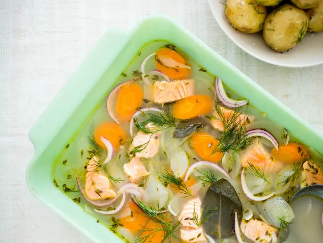 En ljusgrön form med inkokt lax, morötter, lök och dill. En skål med kokta potatisar syns i bakgrunden.