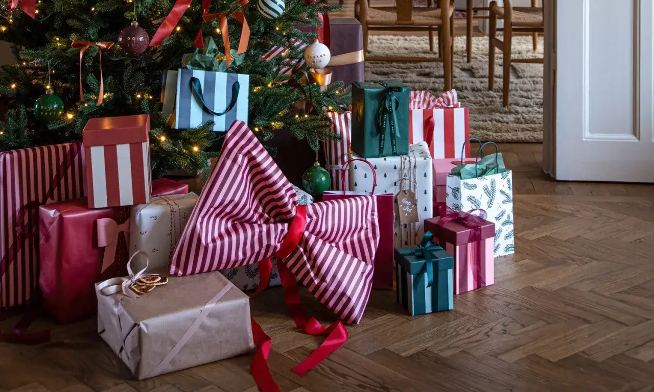 Julklappar i olika färger och mönster ligger under en julgran. En stor rosett av randigt papper dominerar.
