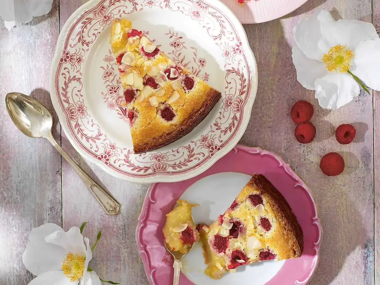 En bit vit kladdkaka med hallon och mandel på en vacker tallrik. En sked med en bit av kakan ligger bredvid.